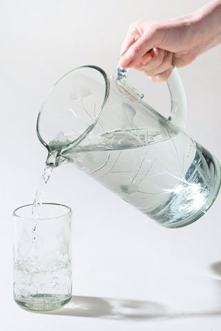 Clear glass pitcher being filled with water from a glass container on a light background