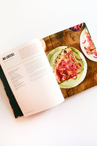 Open cookbook showing a pizza recipe with a wooden cutting board and pizza slice on a white background